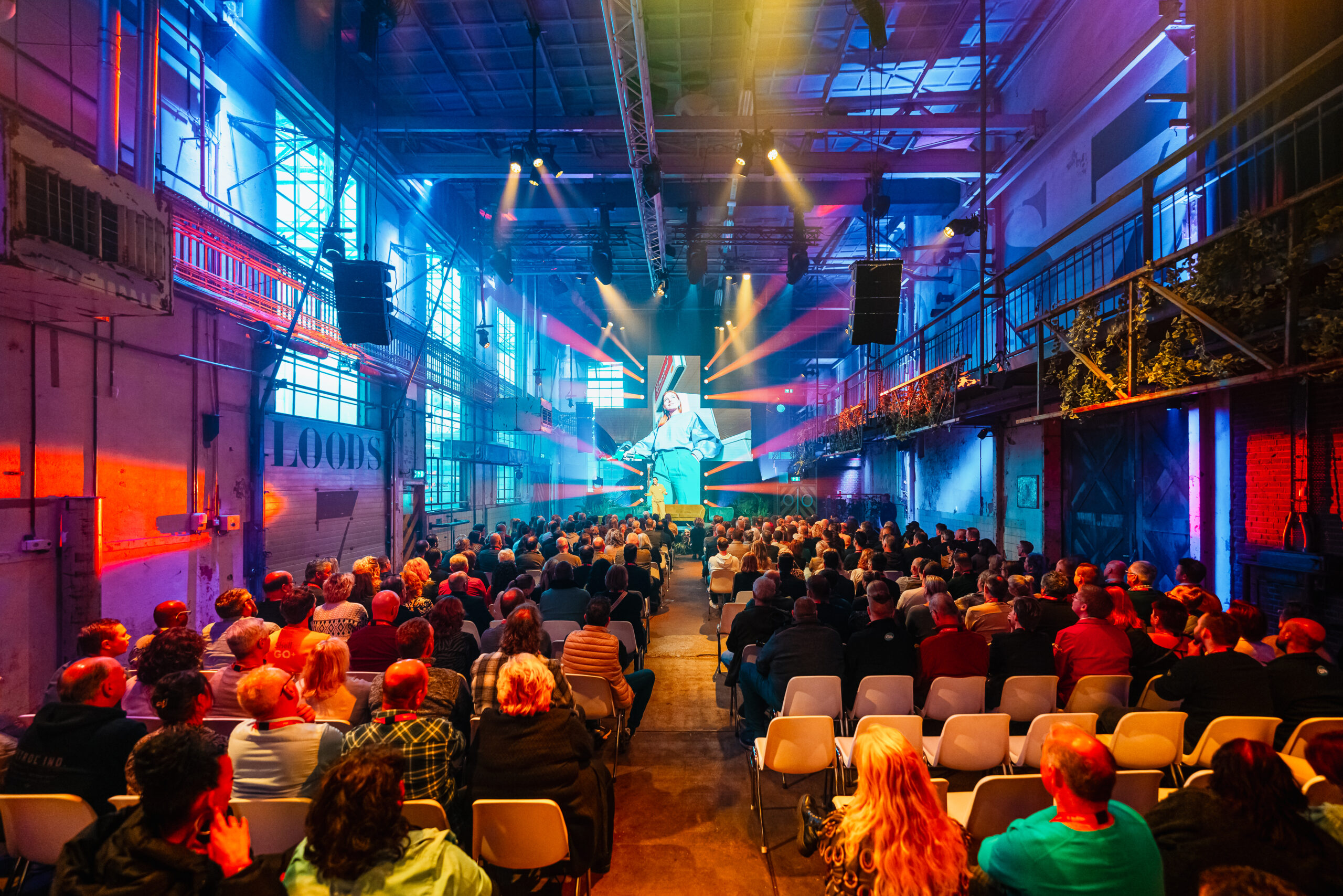 Bedrijfscongres - Publiek in een industriële loods kijkt naar een verlichte presentatie op een groot scherm, met kleurrijke lichteffecten en een podium in het midden.