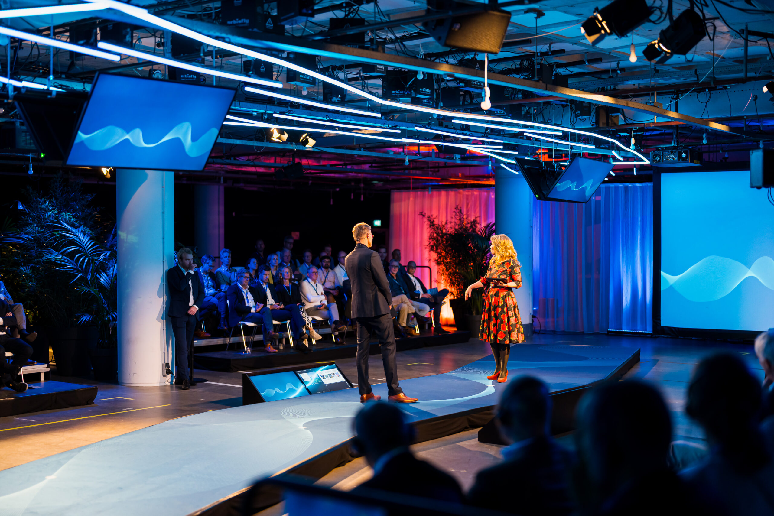 Een professionele presentatie op een modern podium met sfeervolle verlichting, waarbij een man en een vrouw het publiek toespreken.