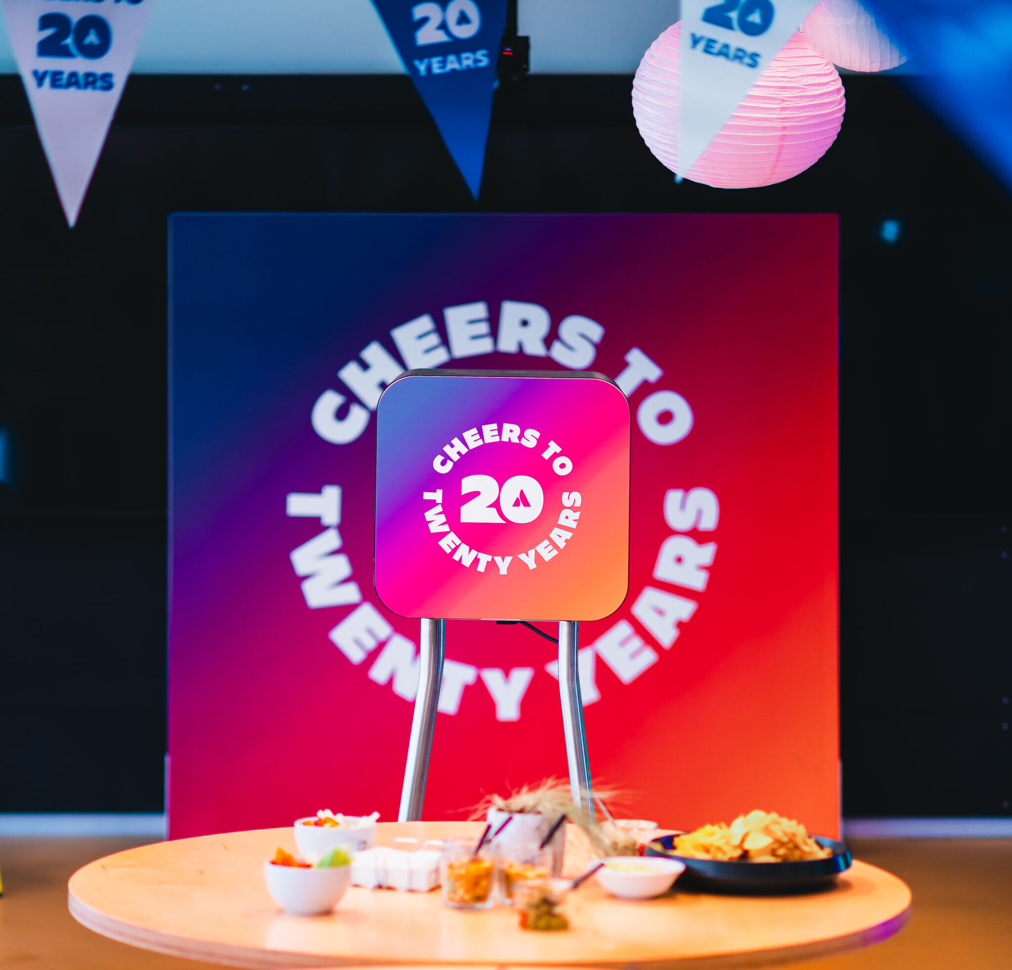 Feestelijke setting met een kleurrijke display met de tekst "CHEERS TO 20 YEARS", decoratieve vlaggetjes en een tafel met hapjes op de voorgrond.