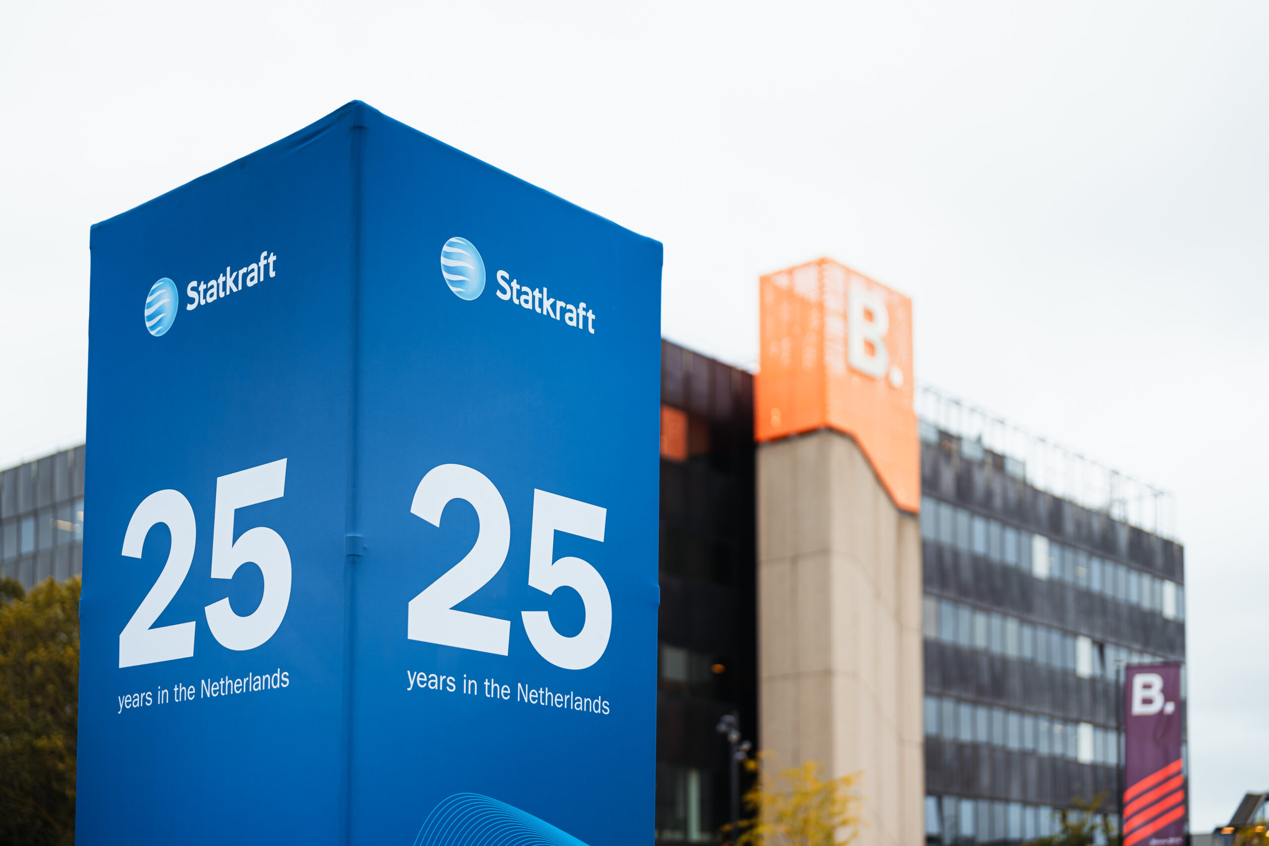 Bedrijfsjubileum - Een grote blauwe banner met het logo van Statkraft en de tekst "25 years in the Netherlands" staat voor een modern kantoorgebouw met een oranje "B."-logo op de gevel. Uitnodigingstraject van je evenement