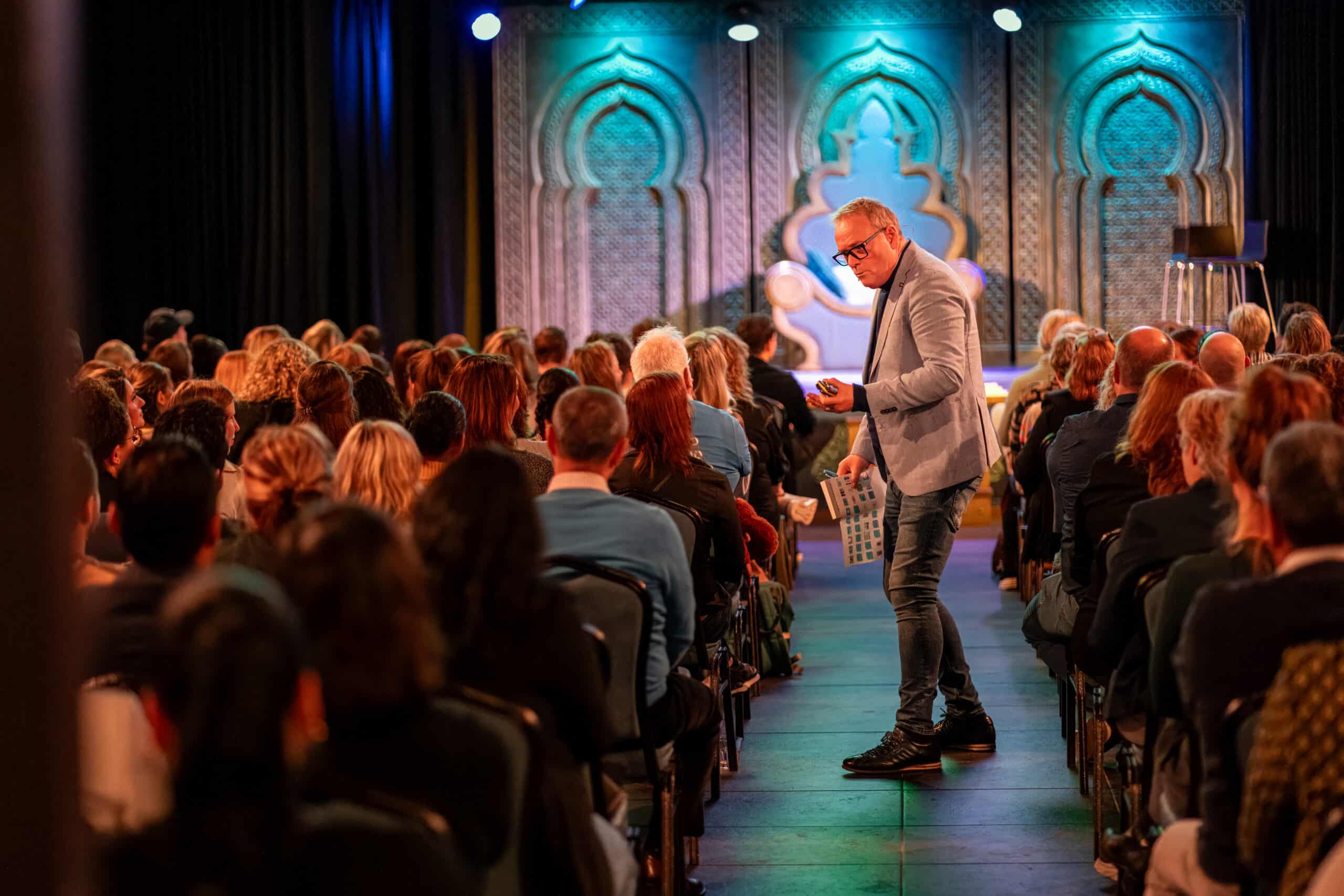 A speaker in a light blue jacket moves down the aisle of a packed auditorium during a conference, with an attractively lit stage in the background.