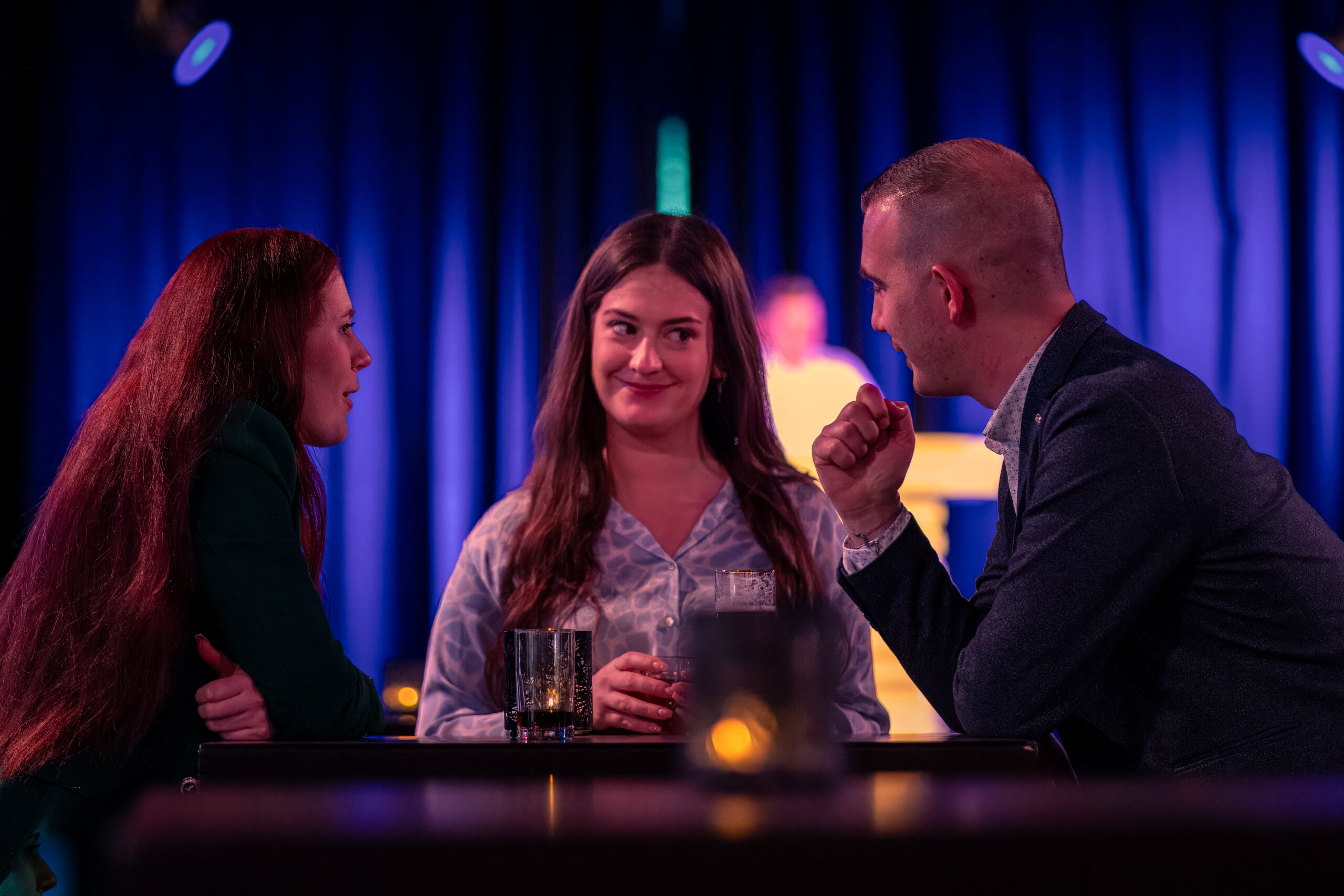 Drie mensen in een sfeervol verlichte ruimte met een blauwe achtergrond voeren een geanimeerd gesprek tijdens een netwerkbijeenkomst.