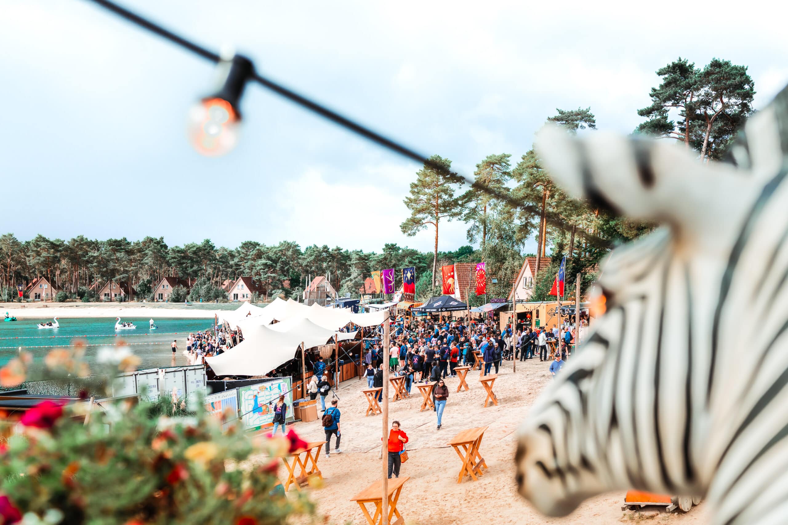 Een levendig bedrijfsfestival organiseren aan een meer met mensen die genieten van eten, drinken en entertainment, terwijl kleurrijke vlaggen en sfeervolle decoratie de omgeving verfraaien.