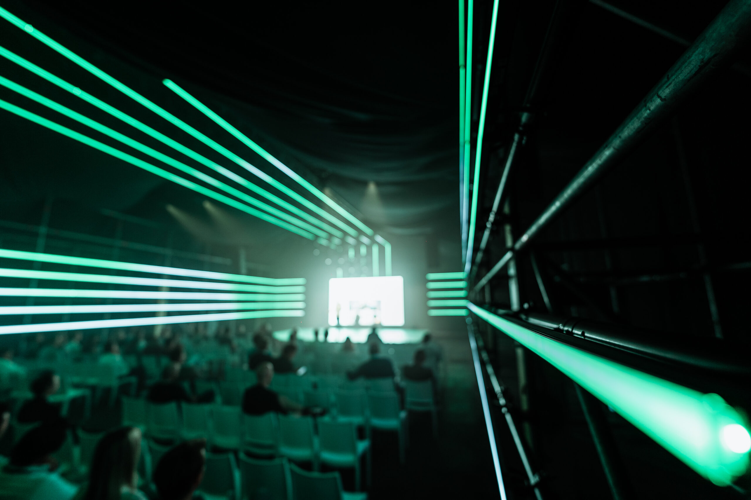 Techniek bij evenement met groene LED-lichtlijnen die naar een podium met een groot scherm leiden, terwijl een publiek op stoelen toekijkt.