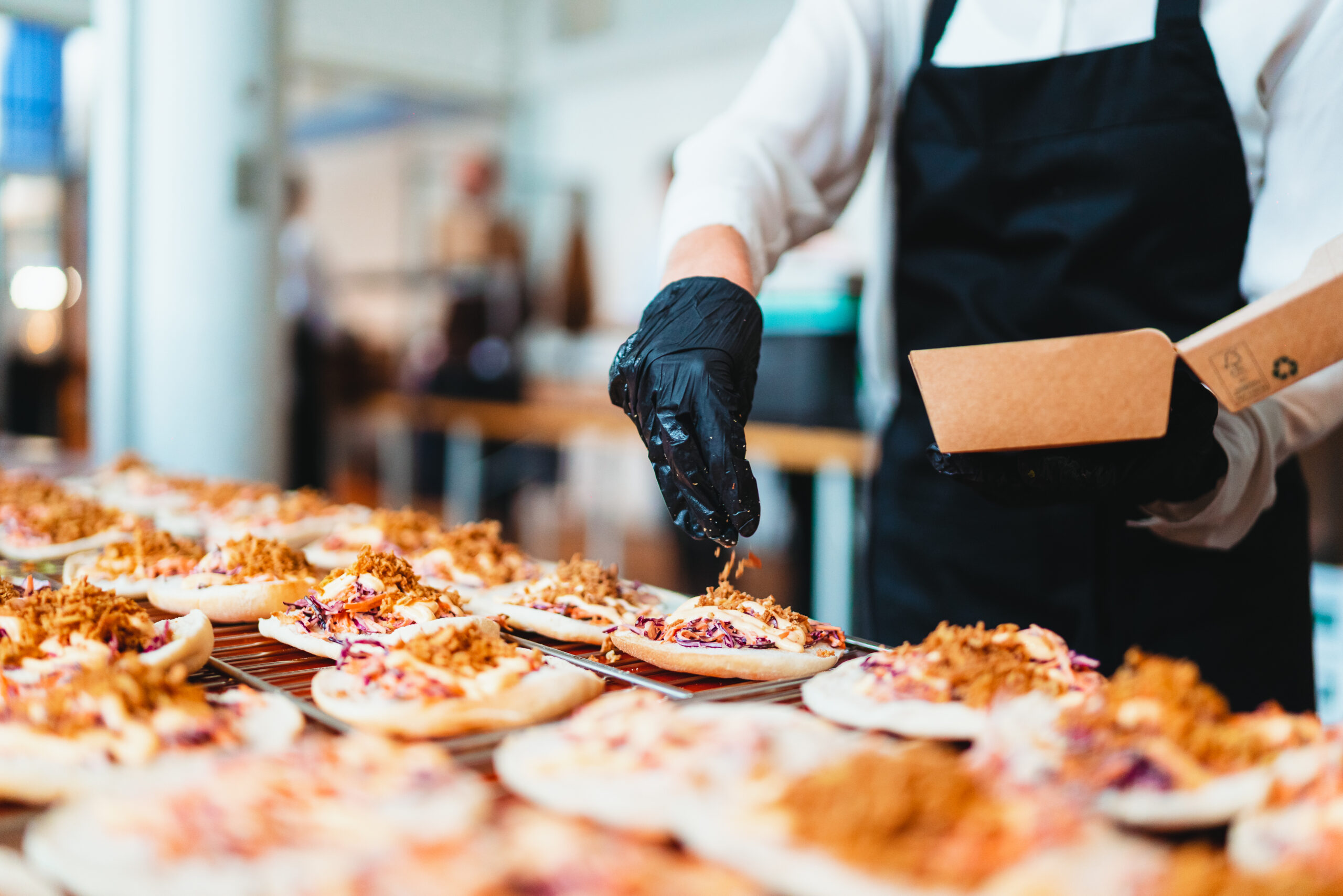 Een cateraar met zwarte handschoenen en een schort bereidt en garneert kleine flatbread-gerechten met knapperige toppings tijdens een evenement.