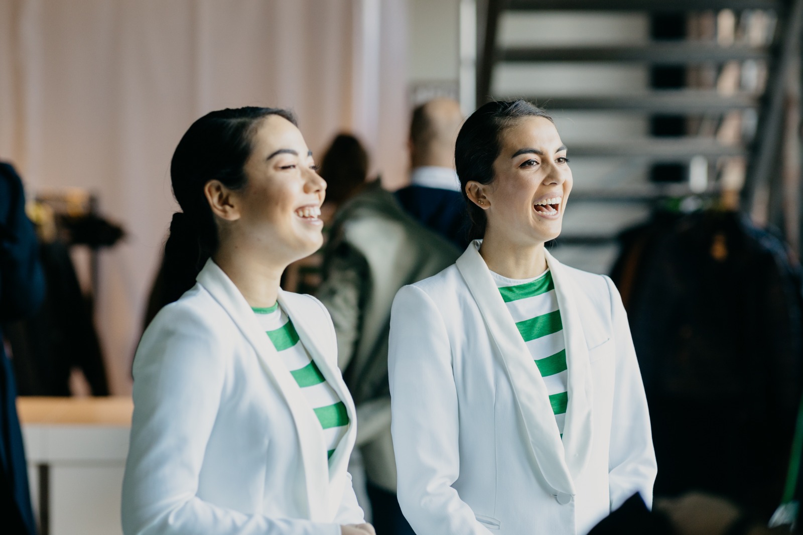 Twee identiek geklede vrouwen in witte blazers en groen-wit gestreepte shirts lachen en praten in een zakelijke evenementenruimte.