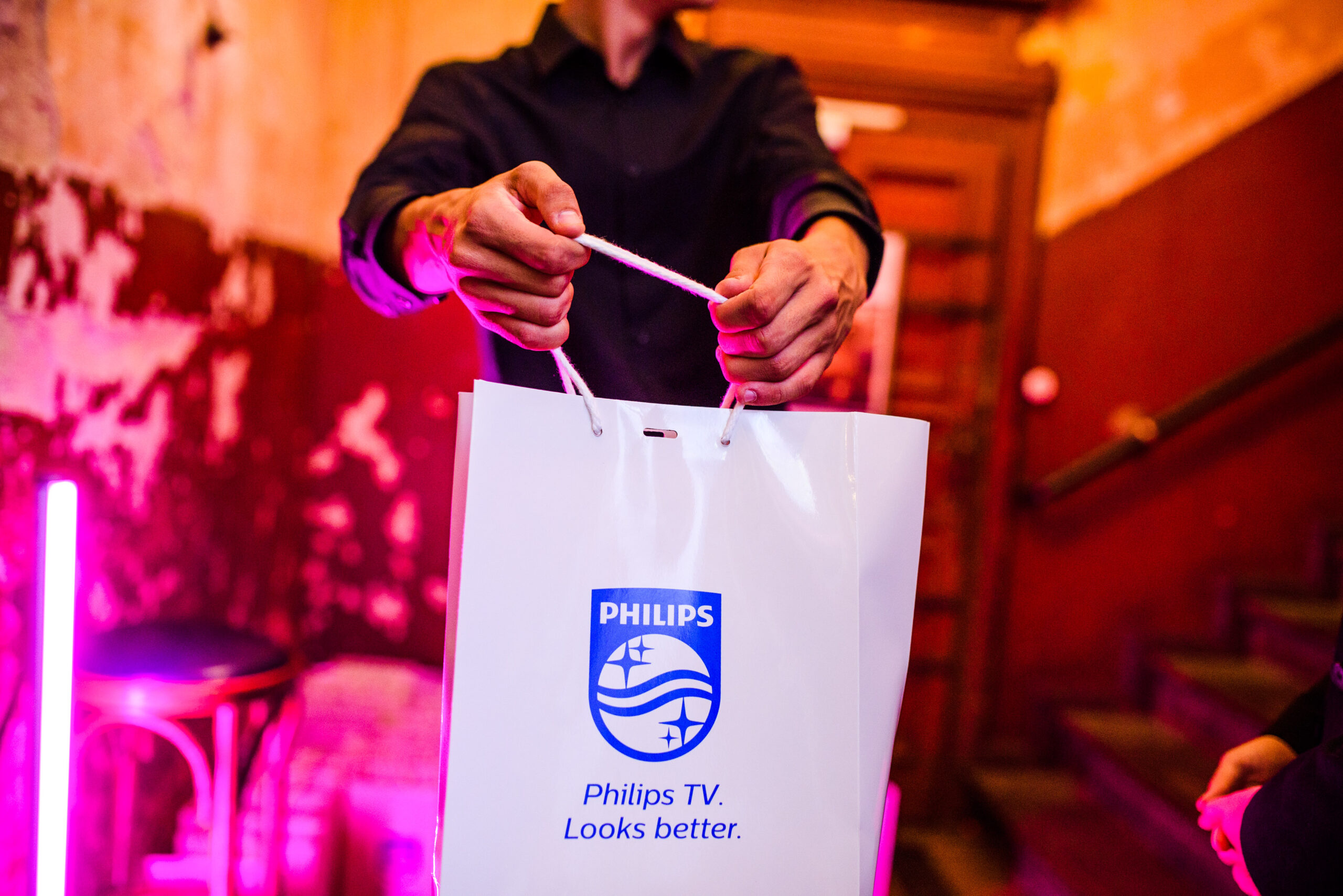 Handen van een persoon in zwart overhemd die een Philips-goodiebag vasthoudt met de tekst 'Philips TV. Looks better.