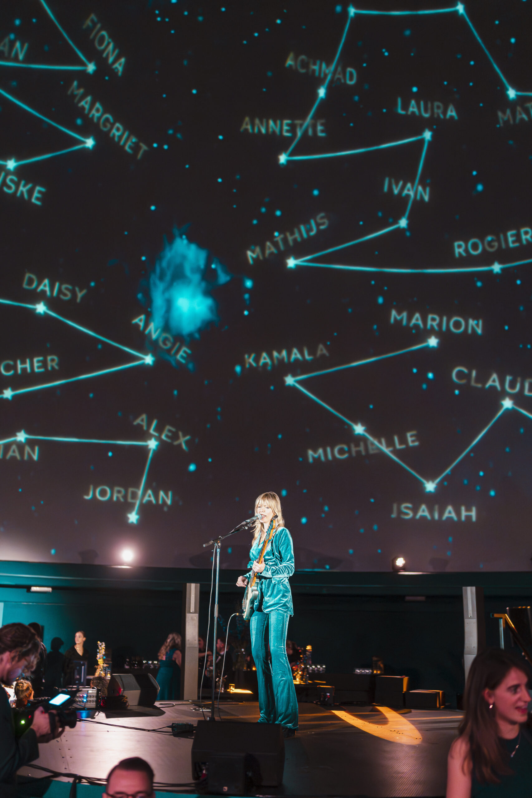 Een muzikant in een turquoise outfit speelt gitaar en zingt op een podium in een futuristische setting. Op de achtergrond is een grote, koepelvormige projectie van een sterrenhemel te zien, met namen verbonden door sterconstellaties.