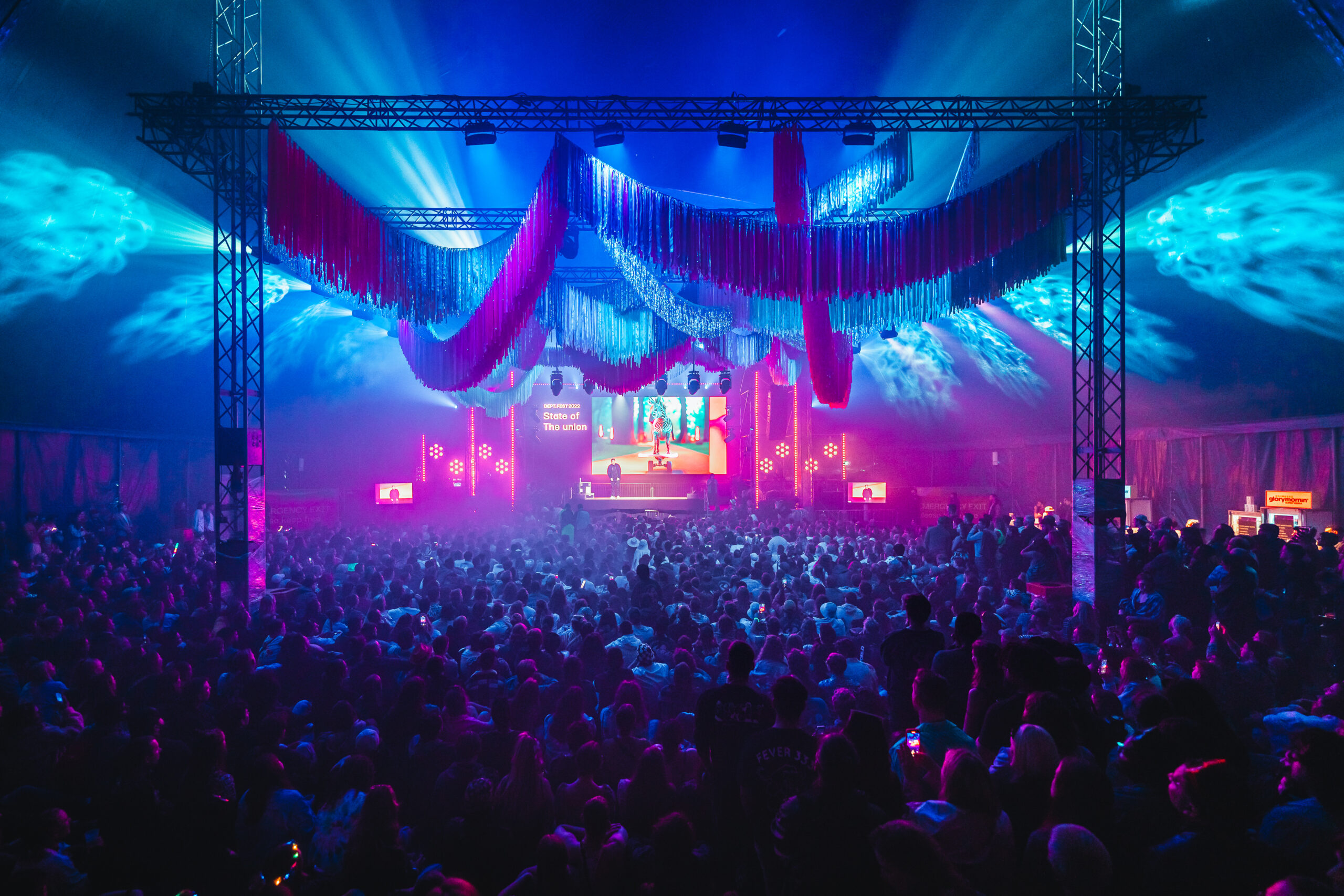 Grootschalig evenement in een sfeervolle tent met kleurrijke verlichting en een podium waar een spreker optreedt.