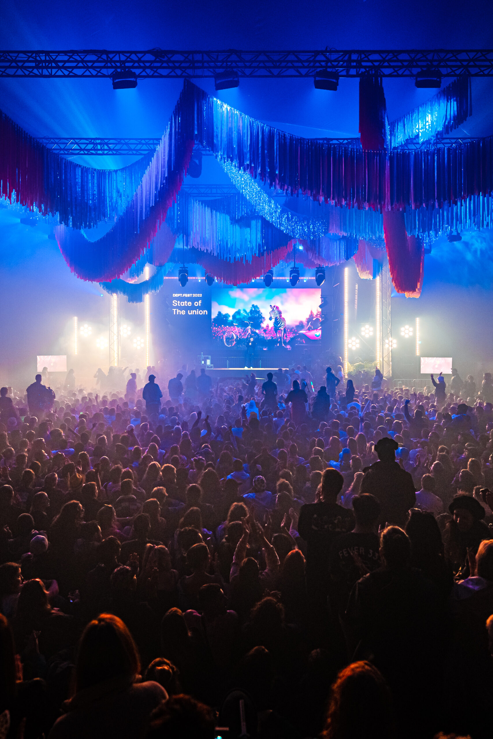 Een volle zaal met een kleurrijke lichtshow, decoratieve franjes en een groot podium tijdens een evenement.